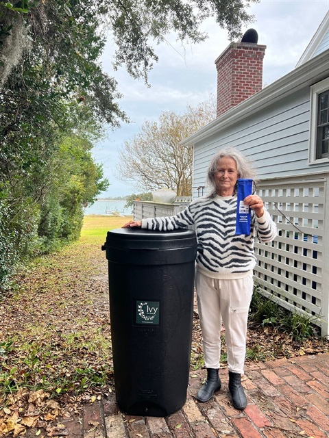 Person standing next to a black rain barrel and holding a Heal Our Waterways giveaway.